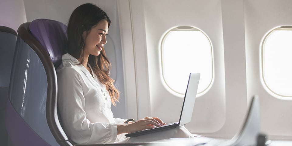 A smiling woman with long hair types on a laptop at her tray table while seated by a sunlit window on an airplane.