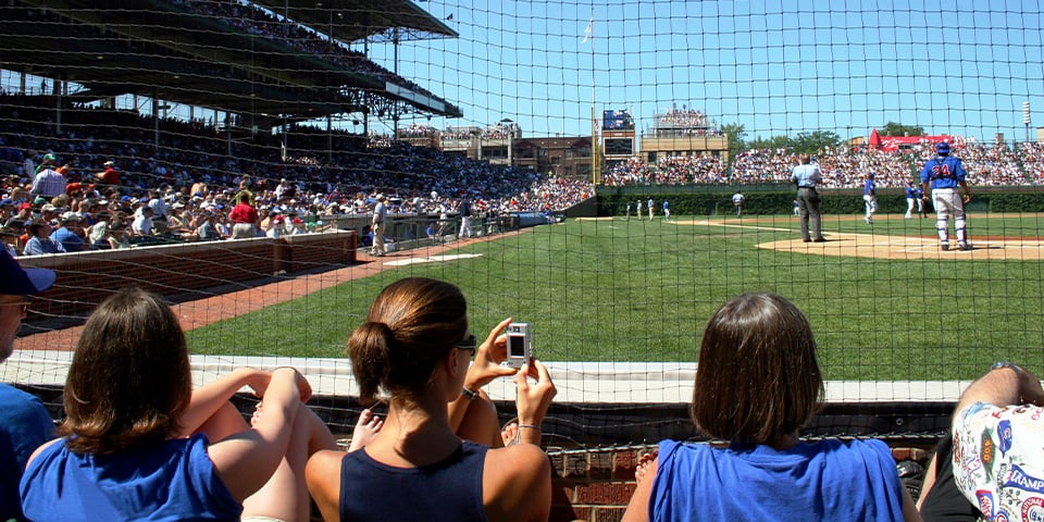 Fans watch a baseball game through protective netting at a packed stadium, one woman taking a photo with a small camera.