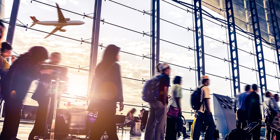 Blurred travelers with luggage move through a sunlit airport terminal as a plane takes off outside the floor-to-ceiling windows.