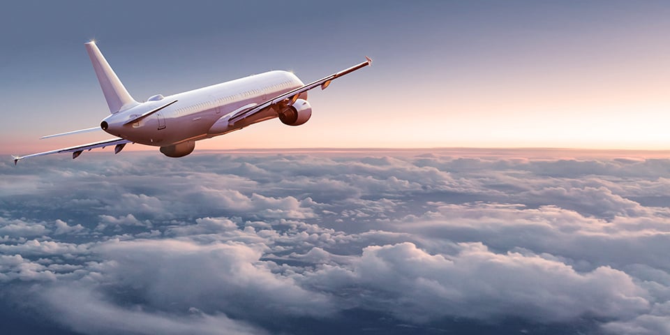 A commercial airplane flies above a thick layer of clouds at sunset, viewed from behind and to the side.