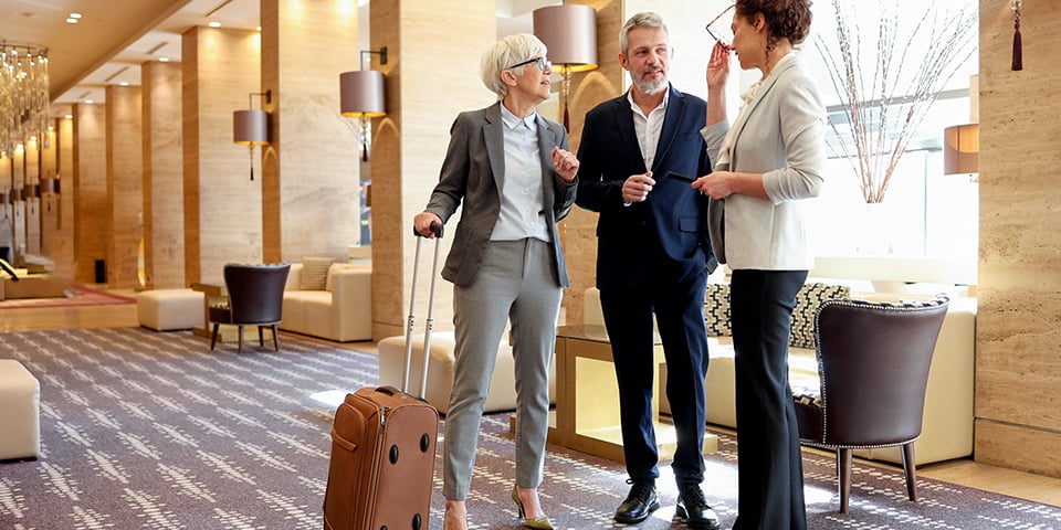 Three business travelers chat in an elegant hotel lobby with marble columns and warm lamps, a woman in a gray suit resting her hand on a brown rolling suitcase as her colleagues talk.
