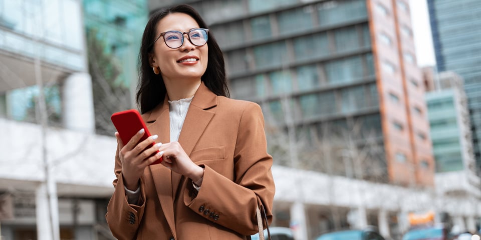 Smiling businesswoman in glasses and a camel blazer holds a red smartphone and looks off to the side while standing on a city street lined with modern office buildings.