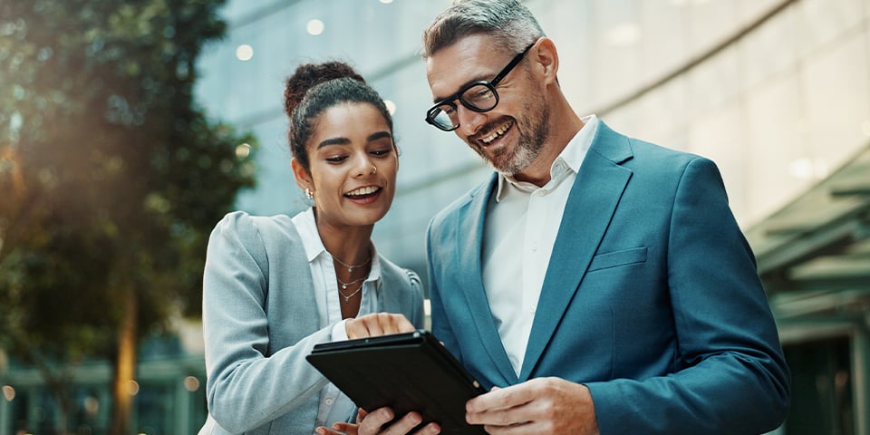 Two smiling colleagues in business attire review a tablet together outside a glass office building, the woman pointing at the screen as a gray-haired man in glasses looks on.