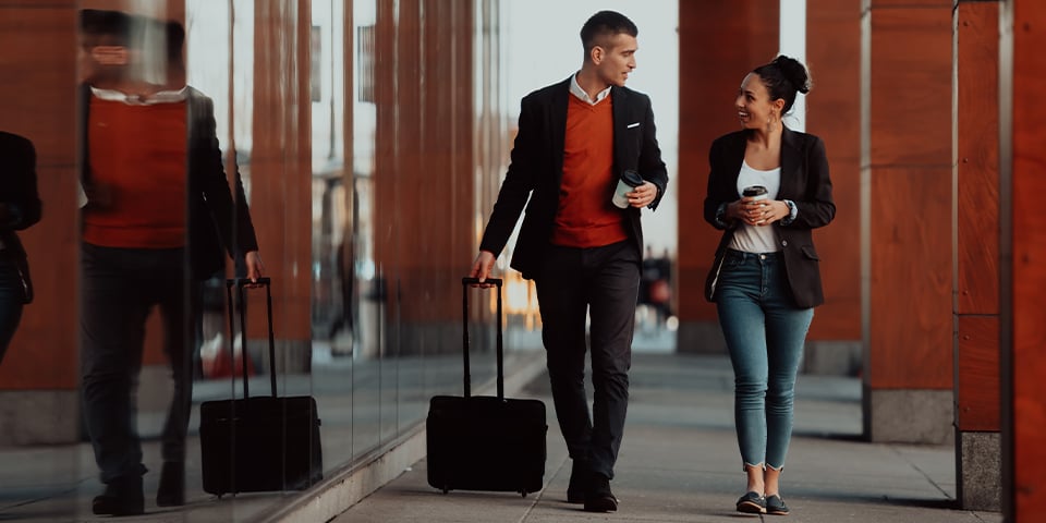 Two smiling business travelers holding coffee cups walk side by side along a glass-walled corridor, the man pulling a black rolling suitcase reflected in the windows beside him.