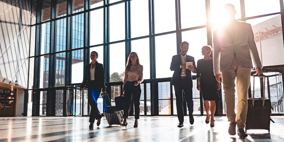 Group of business travelers in professional attire walk with rolling suitcases through a bright, sunlit airport terminal with floor-to-ceiling windows behind them.