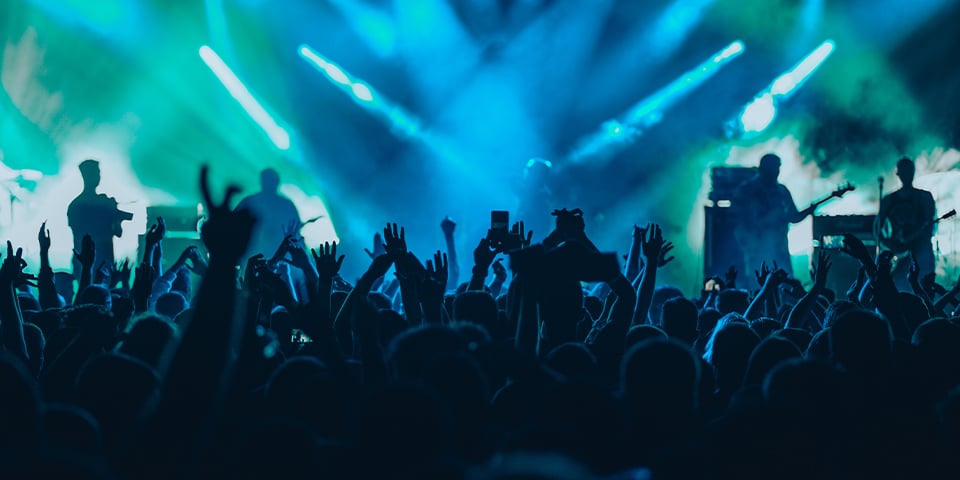 Silhouetted crowd with raised hands at a concert, illuminated by blue and green stage lighting with performers visible on stage.