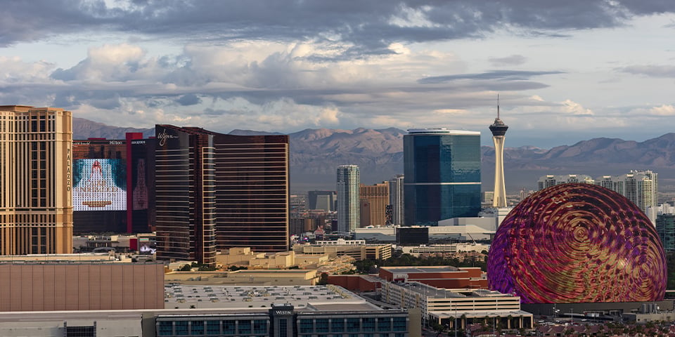 Las Vegas skyline at dusk featuring the Stratosphere Tower, the Sphere, and various hotel and casino buildings against a cloudy sky.