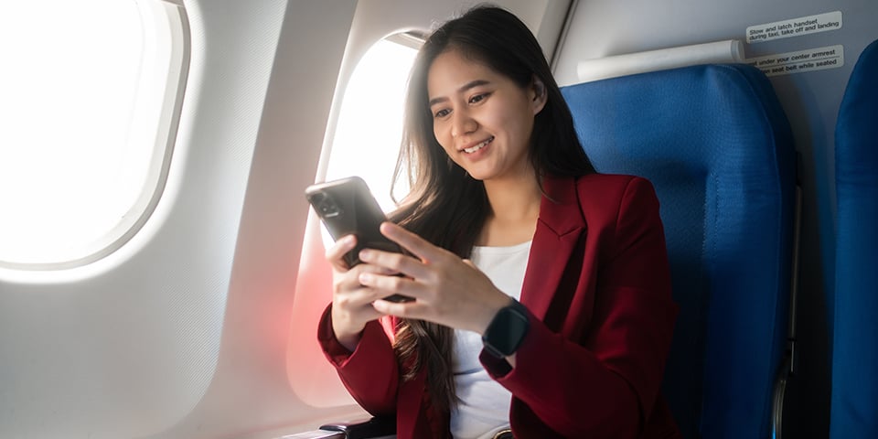 A smiling businesswoman in a red blazer checks her phone while seated in an airplane window seat.