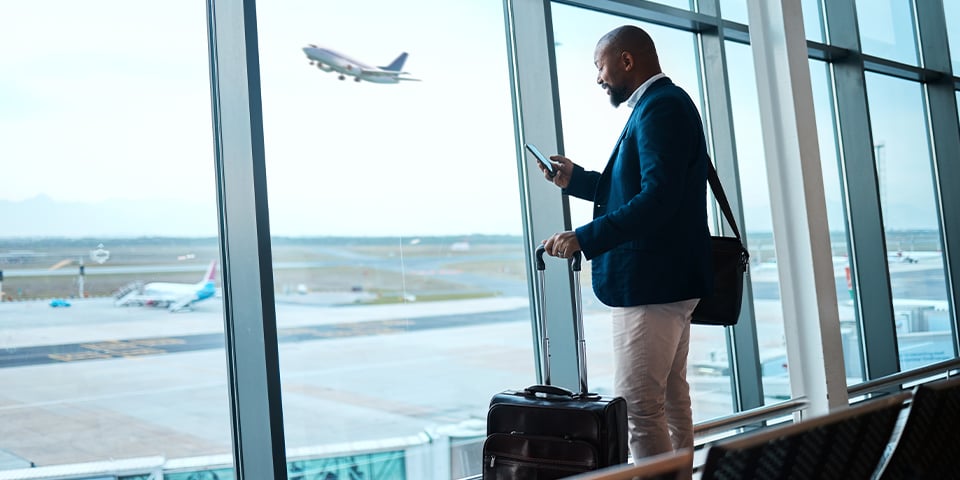 A businessman with a rolling suitcase checks his phone in an airport terminal while a plane takes off on the tarmac outside.