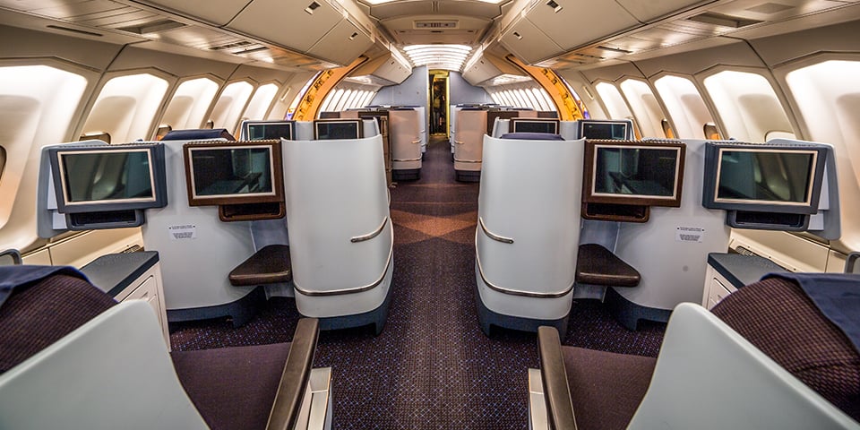 An empty airplane business class cabin with rows of white seats, individual entertainment screens, and warm overhead lighting down the aisle.