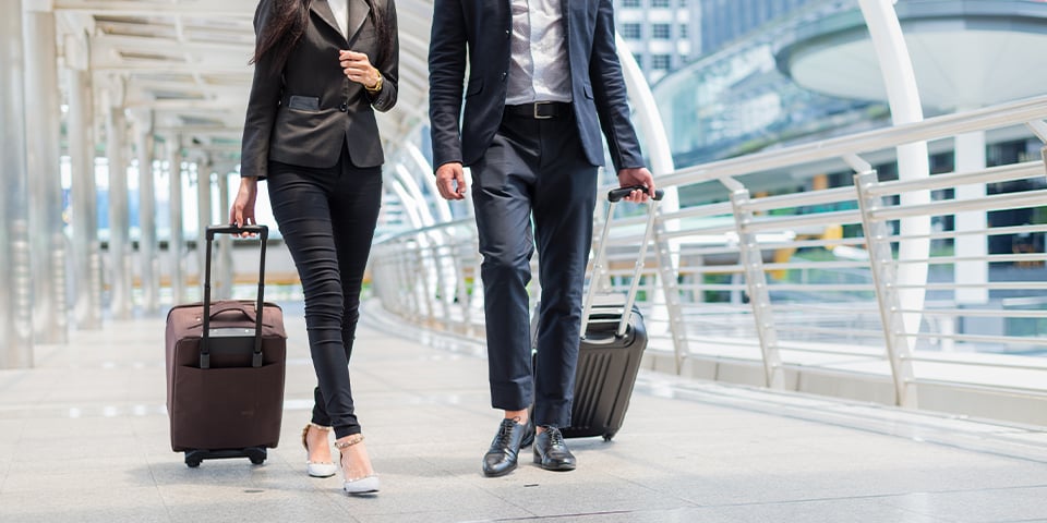 Two business travelers in dark suits walk side by side with rolling suitcases across a bright, covered pedestrian walkway, shown from the shoulders down with modern buildings behind them.