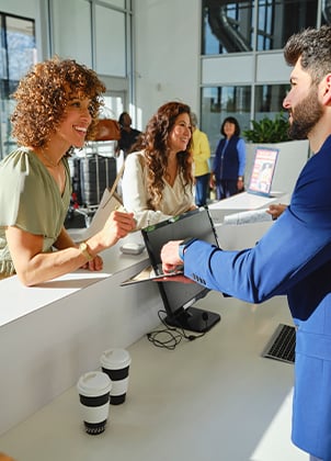 Hotel staff members assist guest with check-in at modern reception desk with coffee cups nearby.