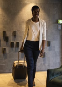 Business traveler in white blazer walks through hotel lobby pulling rolling suitcase.
