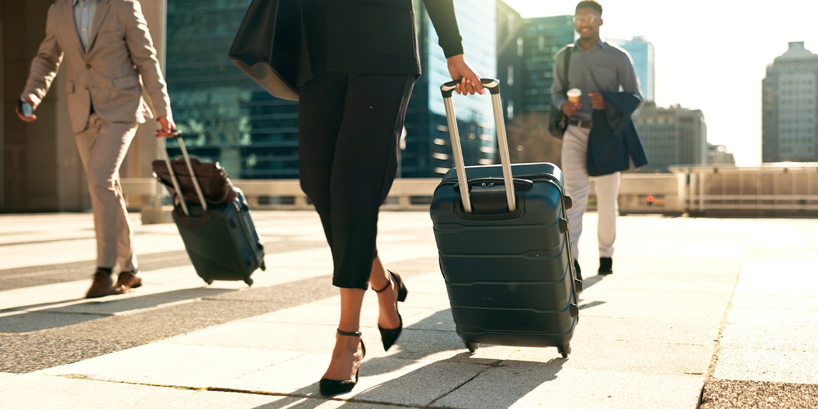 Business travelers walking in bright sunlight with rolling luggage, including a woman in heels in the foreground and two men in professional attire with coffee and bags against an urban skyline backdrop.