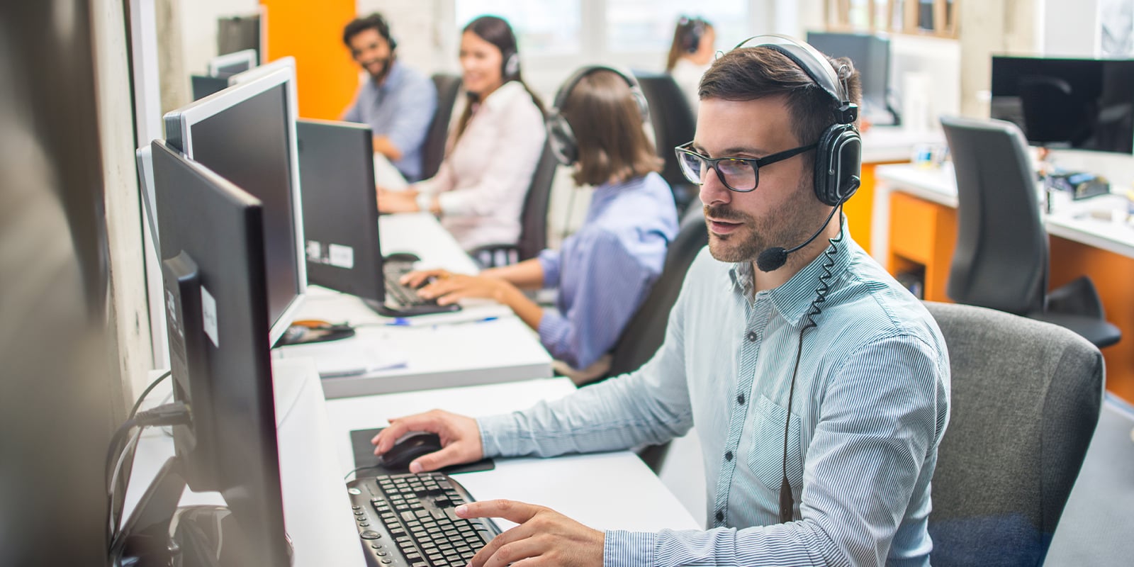 Male customer service representative with headset working in call center office with colleagues in row.