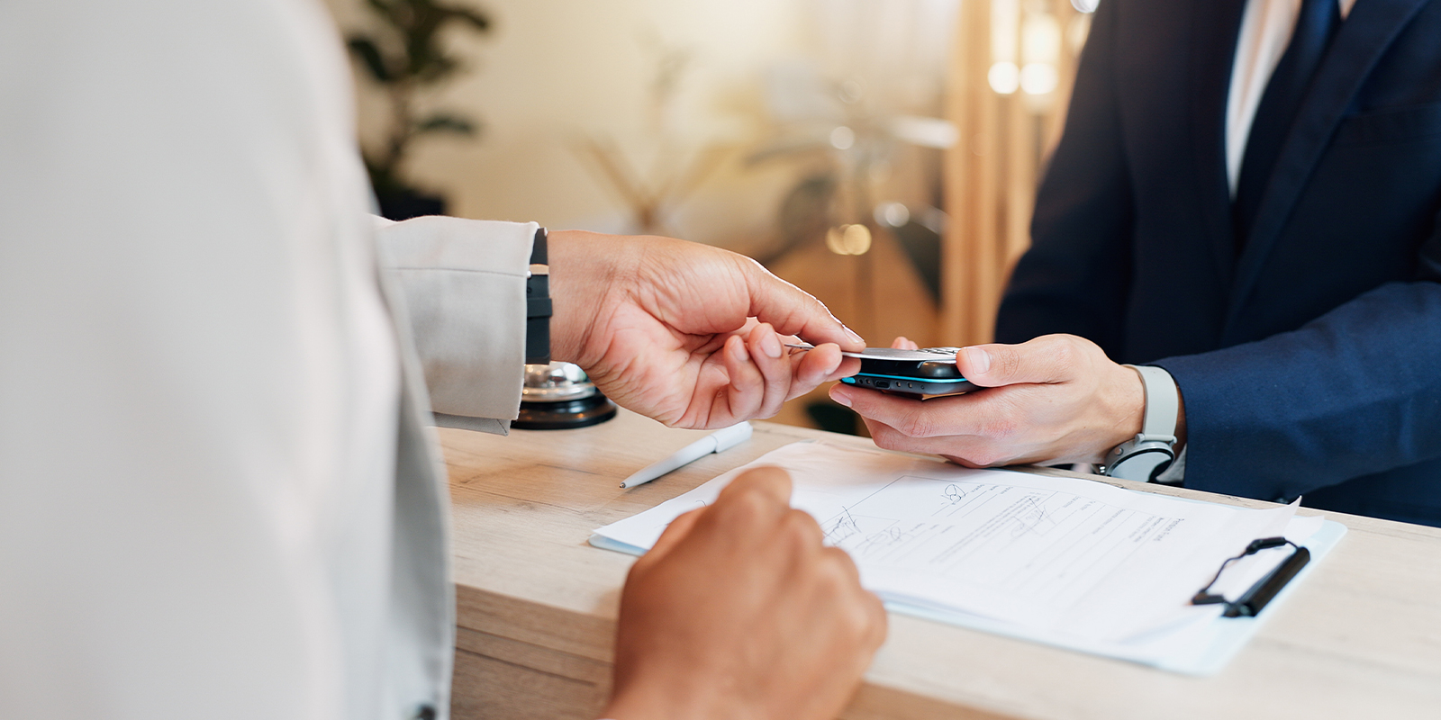 A business professional in a suit using a smartphone while checking in at a hotel reception desk as hotel staff assist with paperwork.