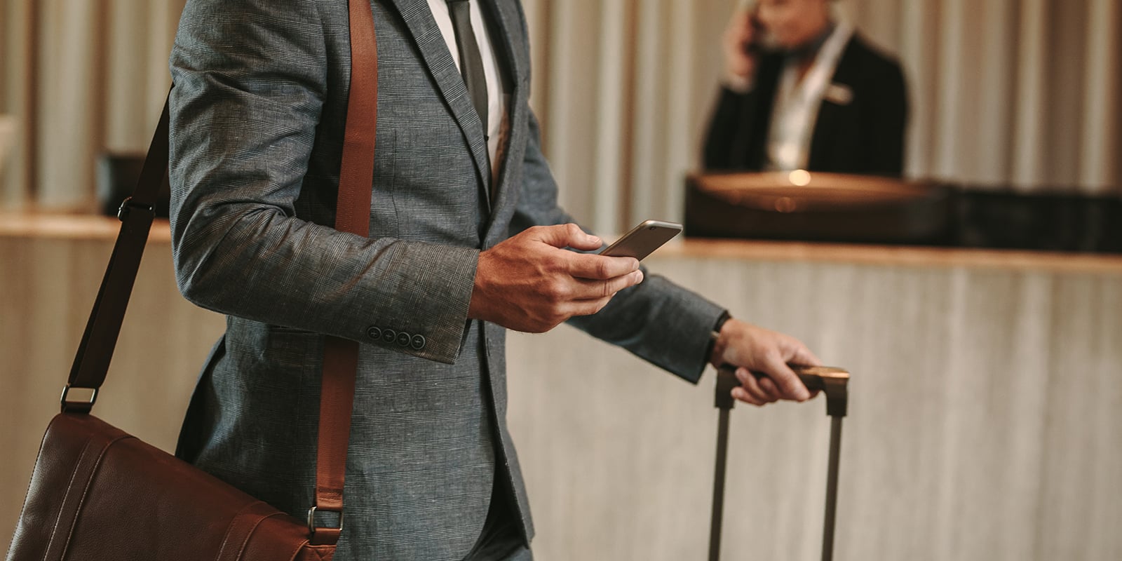 Business traveler in a gray suit with a leather messenger bag checks his smartphone while pulling a rolling suitcase through a hotel lobby, with a receptionist on the phone behind the front desk.